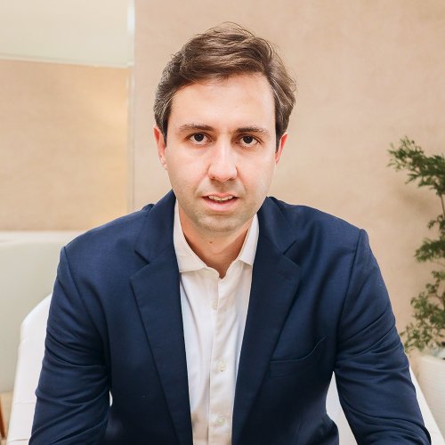 Homem de cabelos castanhos usando terno azul escuro e camisa branca sentado em cadeira branca em ambiente neutro.